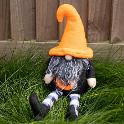 A plush toy resembling a gnome with an orange hat and black outfit, sitting on grass against a wooden fence background.